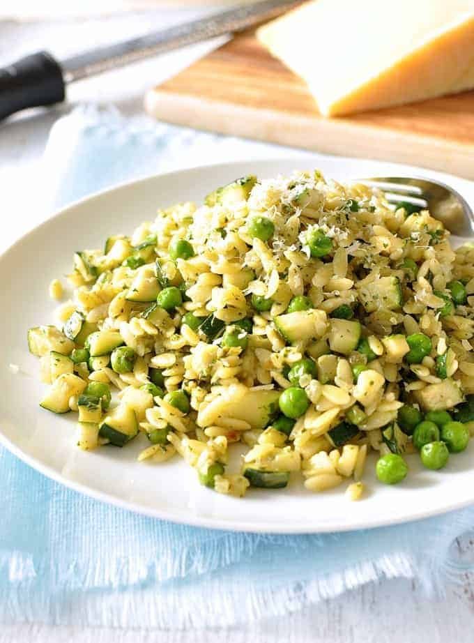 Close up of Pea, Zucchini and Pesto Orzo / Risoni on a white plate with parmesan in the background.